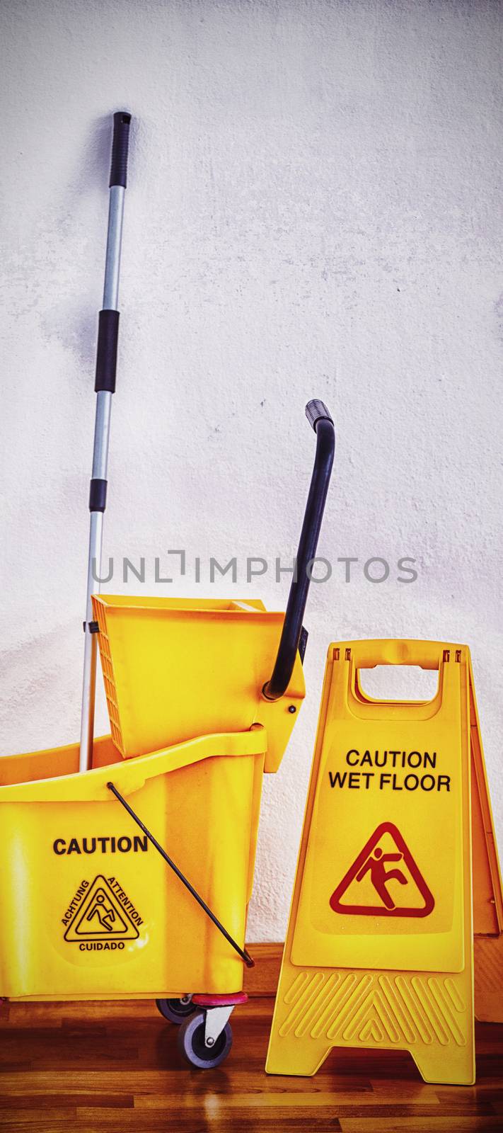 Yellow sign board with mop bucket on wooden floor against wall