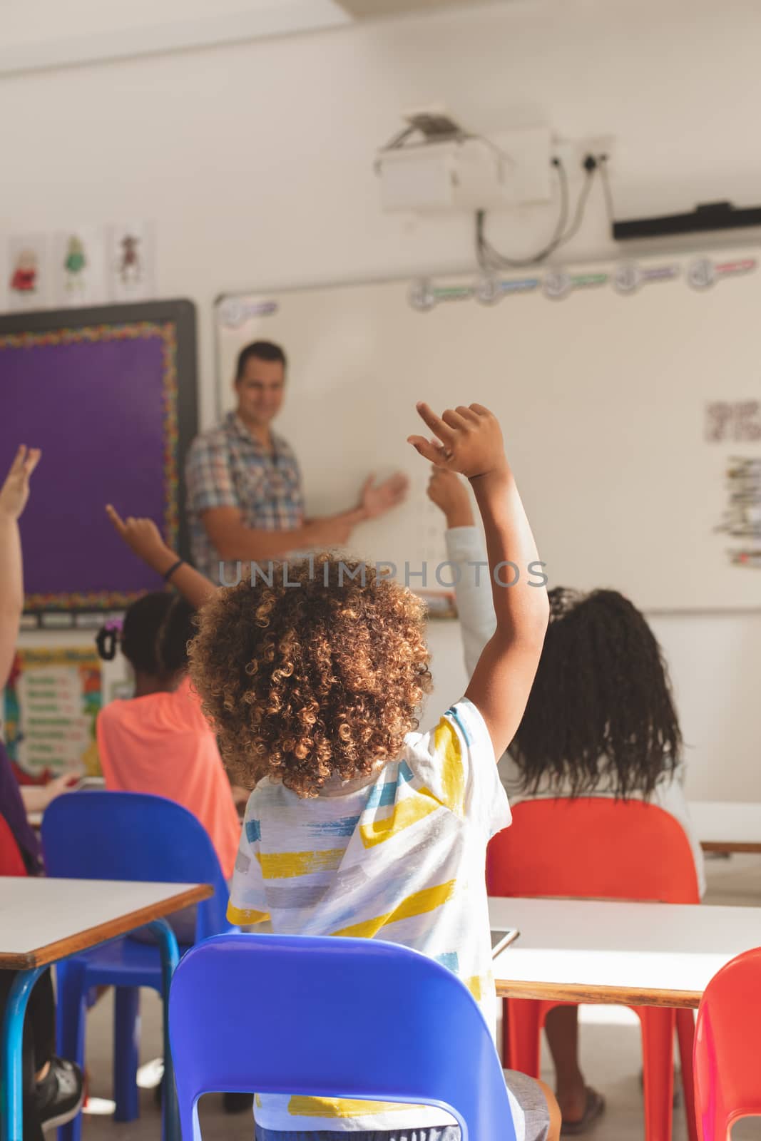 Rear view of school kids raising hand to answer at a question while the teacher speaking at whiteboard