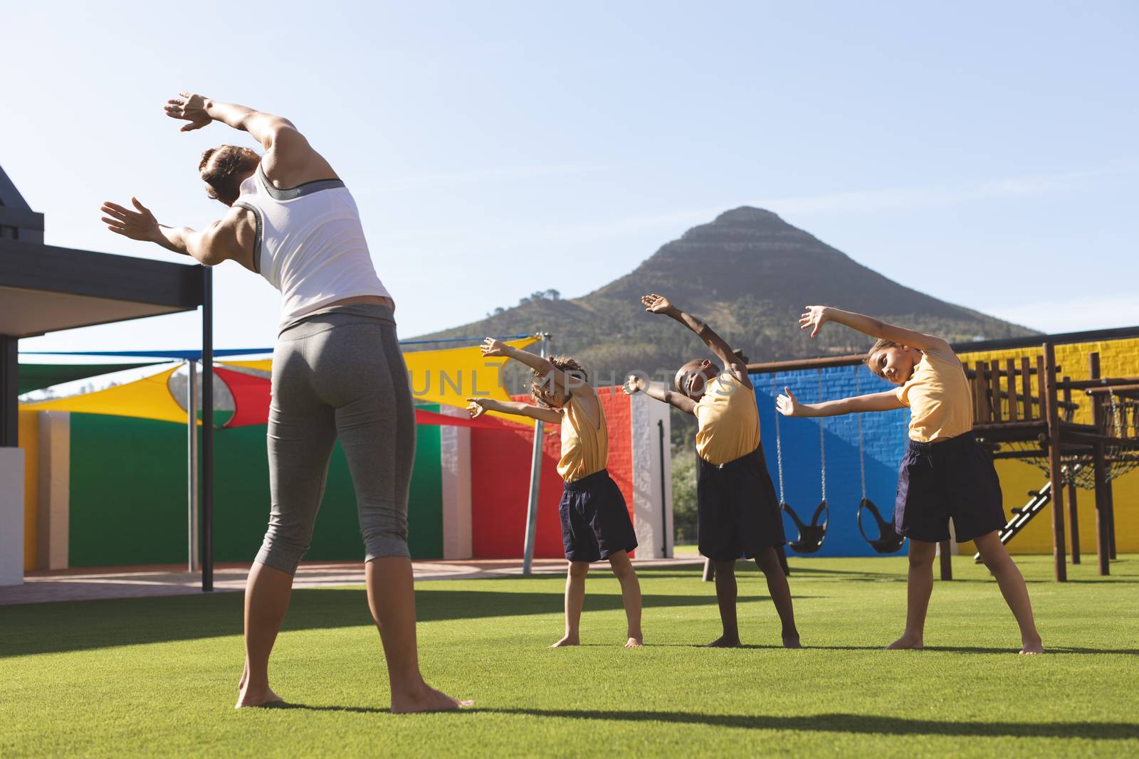 Rear view of caucasian trainer teaching yoga to students in school playground at schoolyard