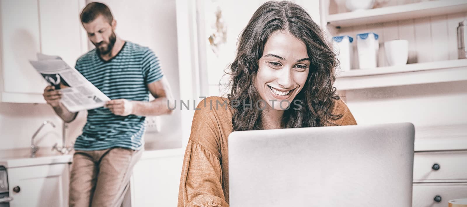 Woman using laptop while man reading newspaper in background at kitchen