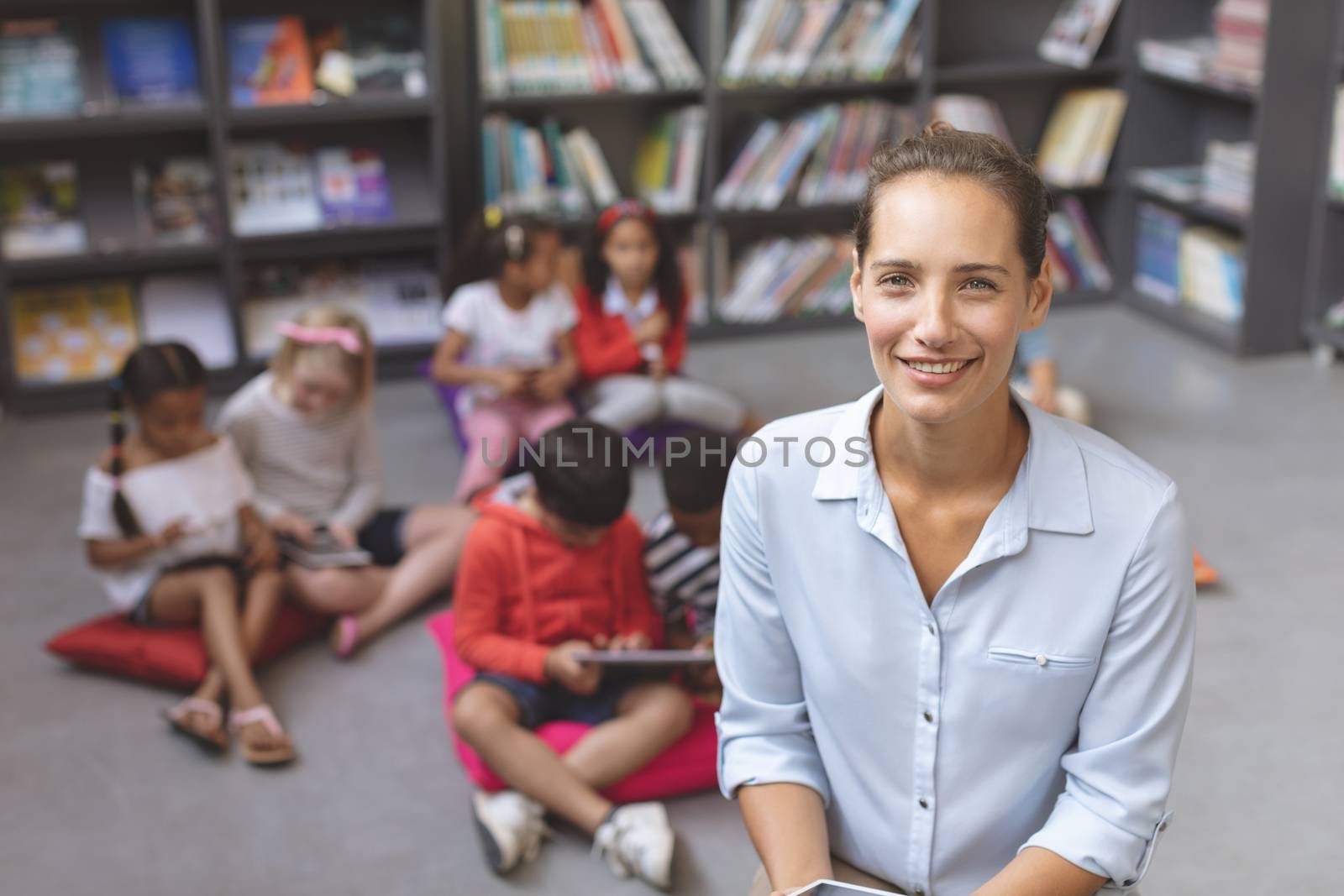 Overhead view of a happy teacher looking and smilling at the camera while school kids relaxing themselves over digital tablet on cushions in background