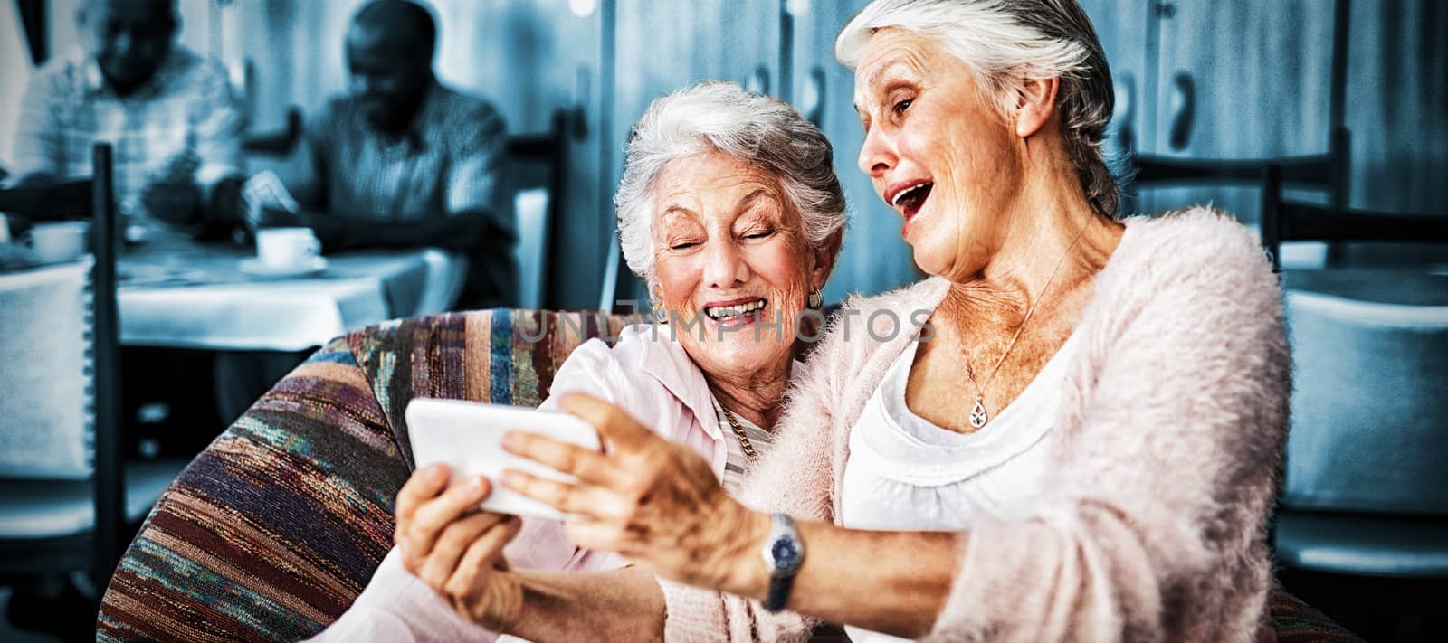 Senior friends taking selfie through smart phone while sitting on sofa at nursing home