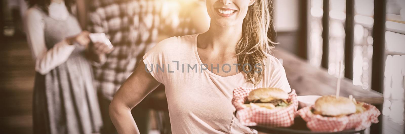 Portrait of smiling young waitress serving burger with customers in background at restaurant