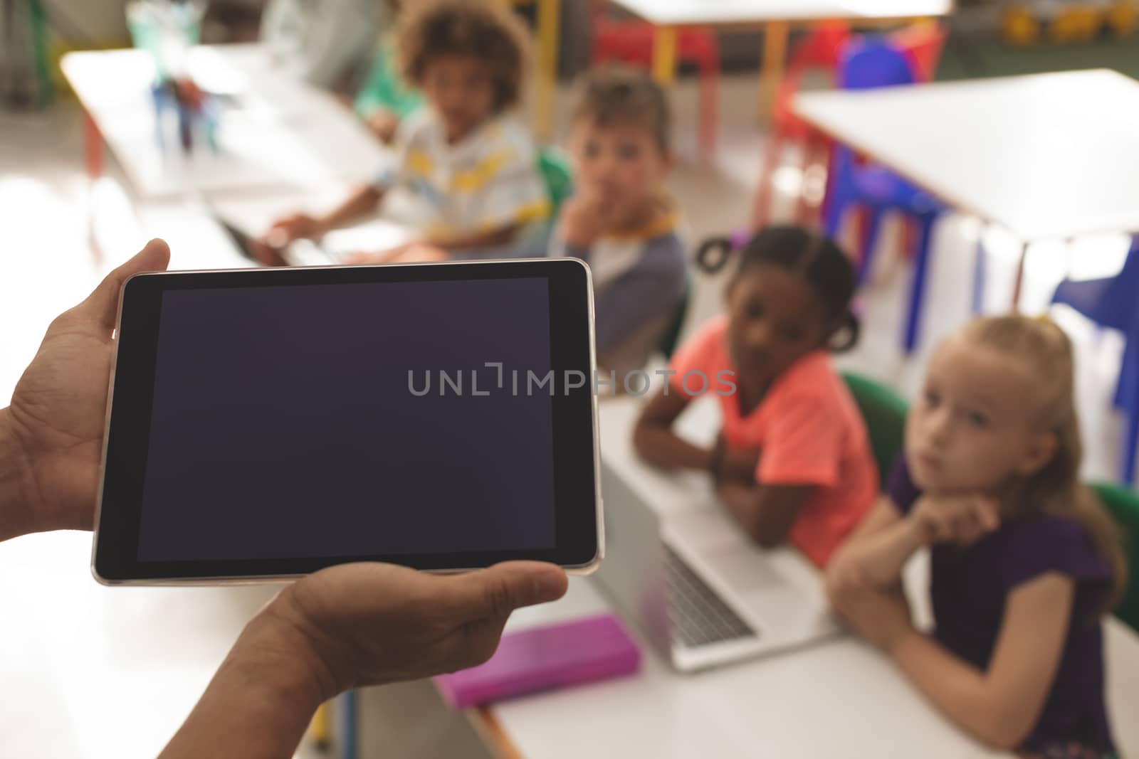 Close-up of a teacher holding a digital tablet with school kids looking at her in classroom at school