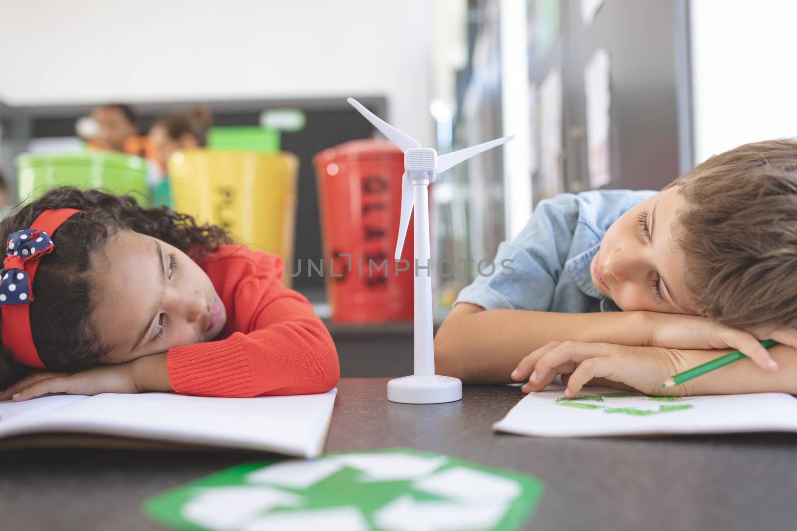 Front view of two tired school kids observing a windmill mockup between them in classroom against trash cans plastic explosives of recycling