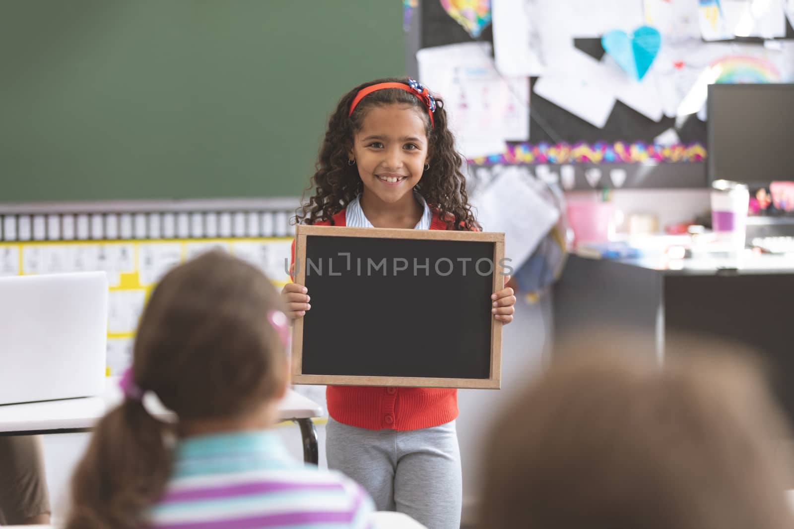 Front view of happy mixed-race schoolgirl holding slate in the classroom at school with his classmates in foreground