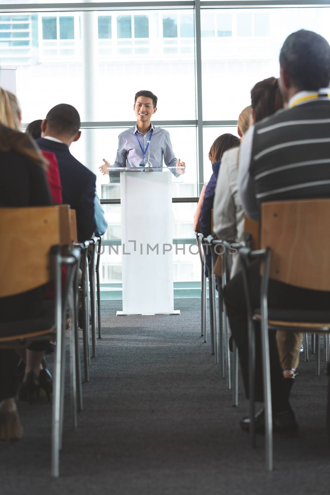 Front view of young Asian businessman smiling as he speaks in front of business professionals sitting at business seminar in office building