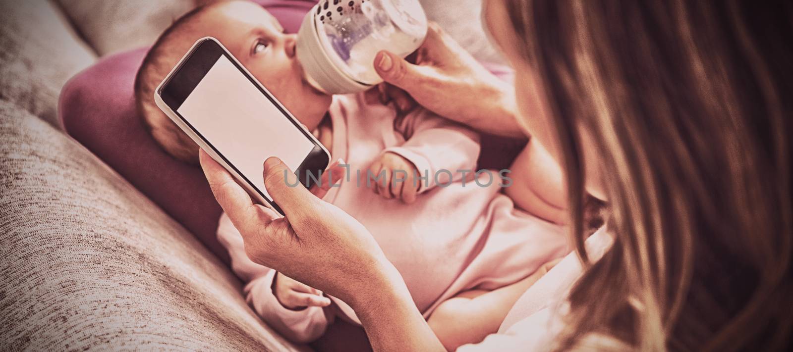 Mother using mobile phone while feeding her baby with milk bottle by Wavebreakmedia