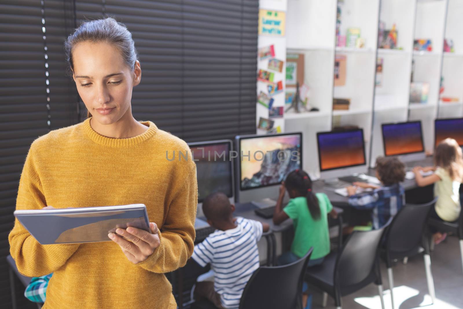 Front view of caucasian teacher using digital tablet while multi ethnic kids working on computer behind her in computer room 