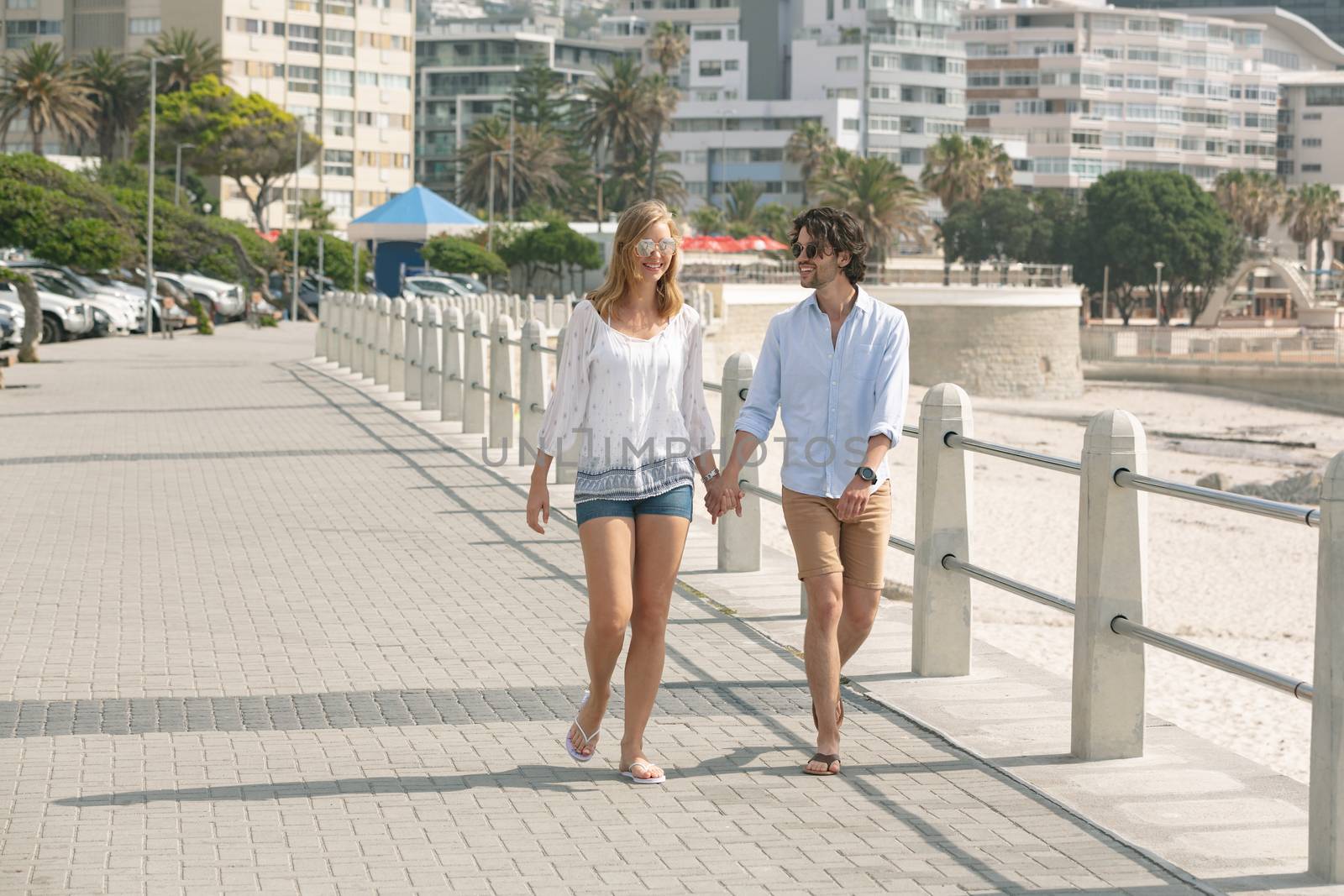 Front view of young Caucasian couple walking hand in hand on the promenade at the seaside on sunny day. They are smiling