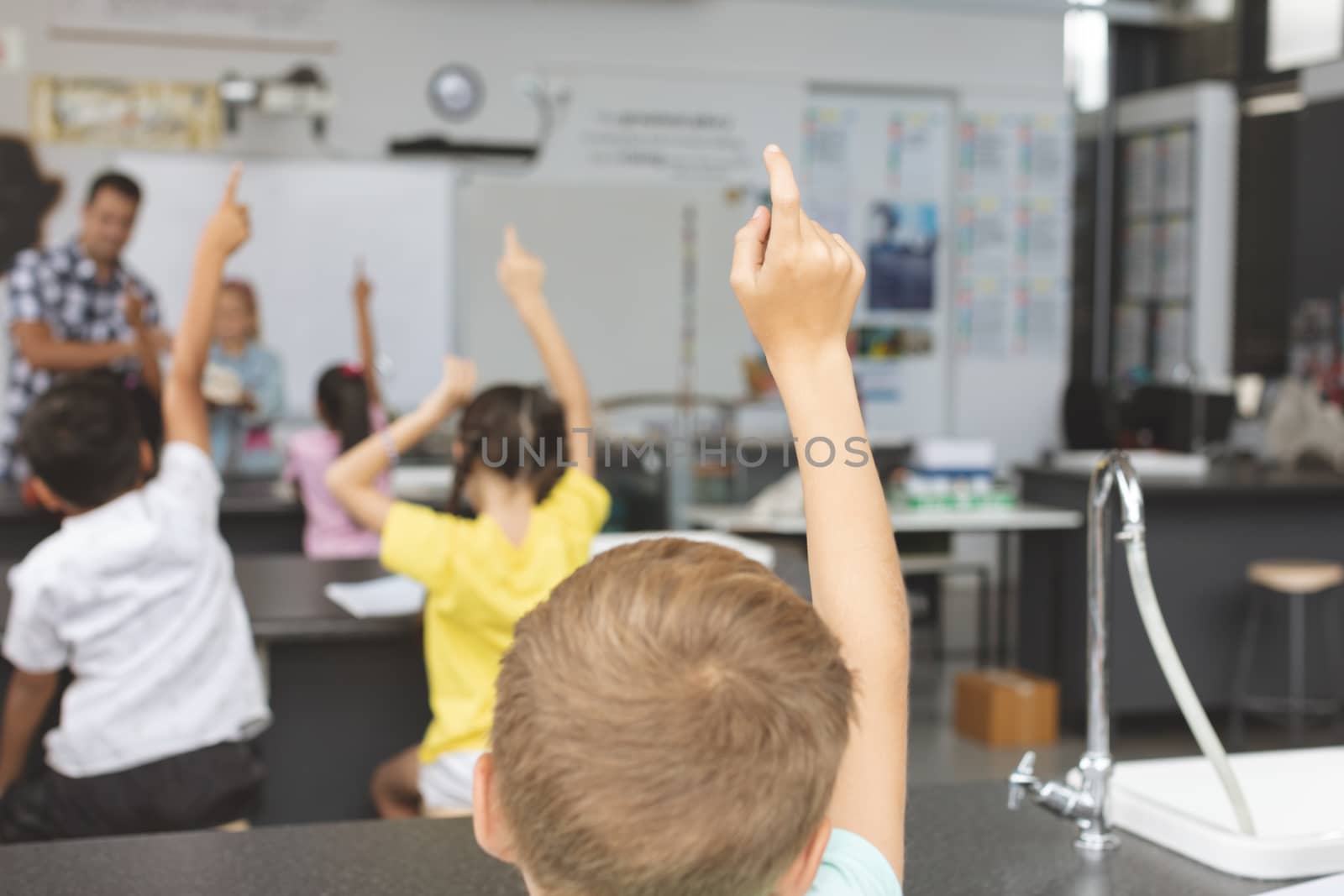 Rear view of school kids raising hand in classroom at school with teacher in background