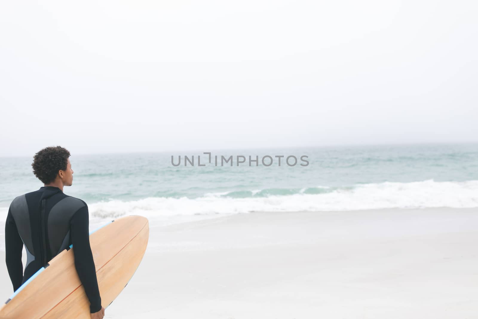 Rear view of thoughtful young mixed-race male surfer holding surfboard on the beach. He is looking away