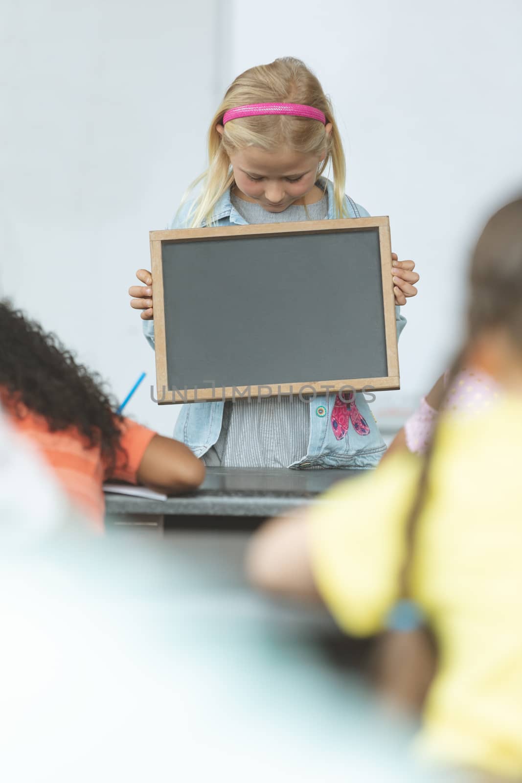 Front view of a Caucasian schoolgirl holding a slate in classroom at school with his classmates sitting in front of her  