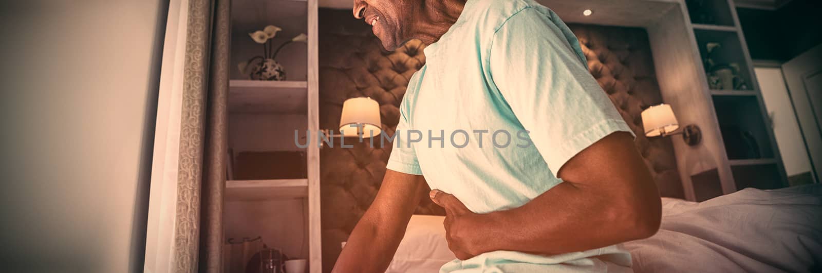 Side view of senior man suffering from stomachache while sitting on bed at home