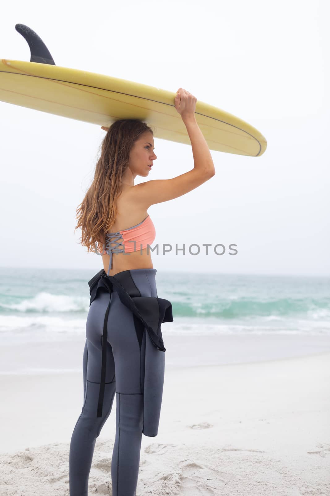 Rear view of beautiful young Caucasian female surfer carrying surfboard on her head at beach. She is looking away