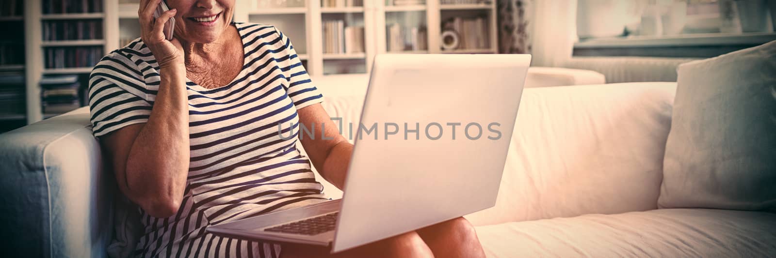 Senior woman talking on mobile phone while using laptop in living room by Wavebreakmedia