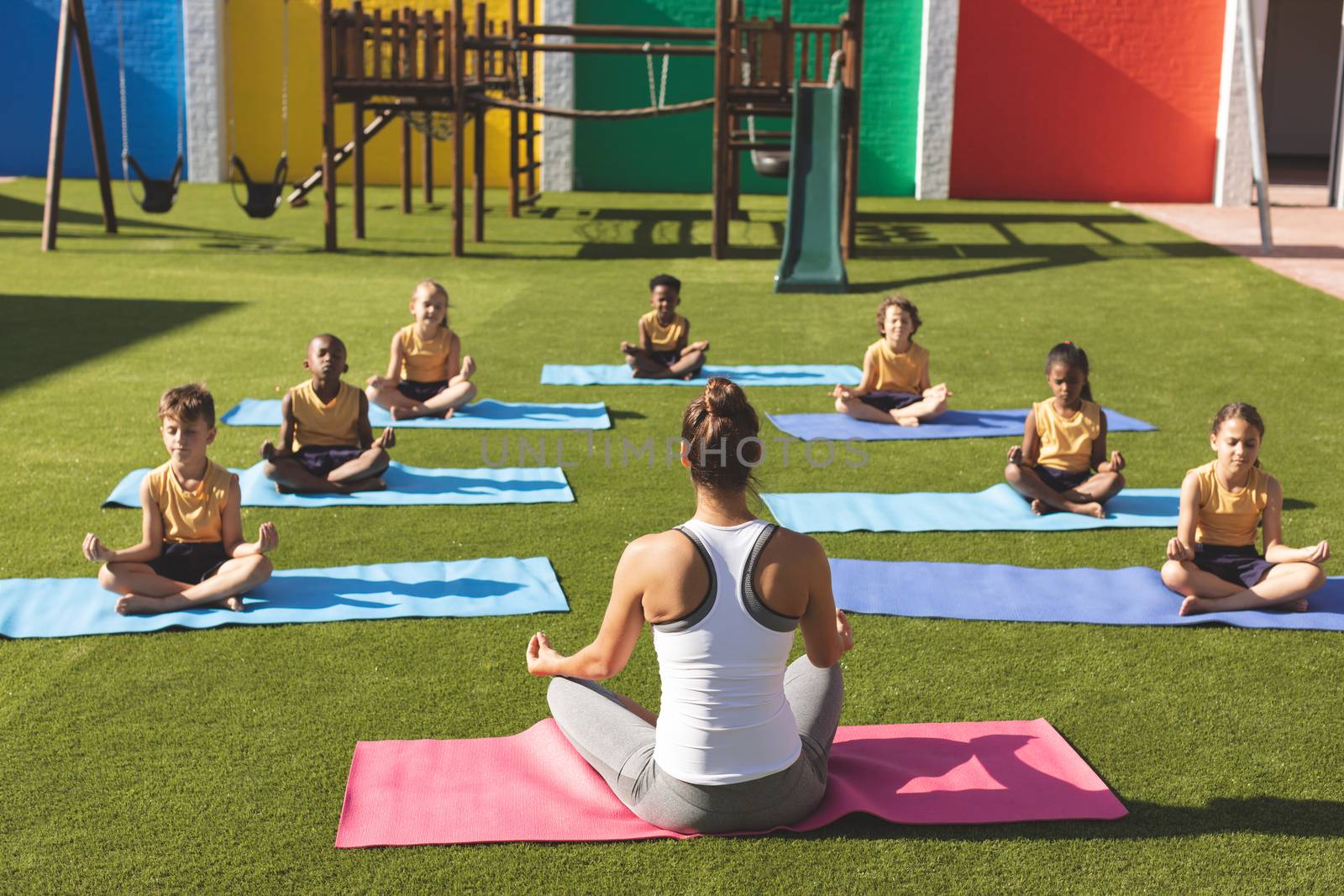 Rear view of caucasian trainer teaching yoga to students on yoga mat in school playground at schoolyard