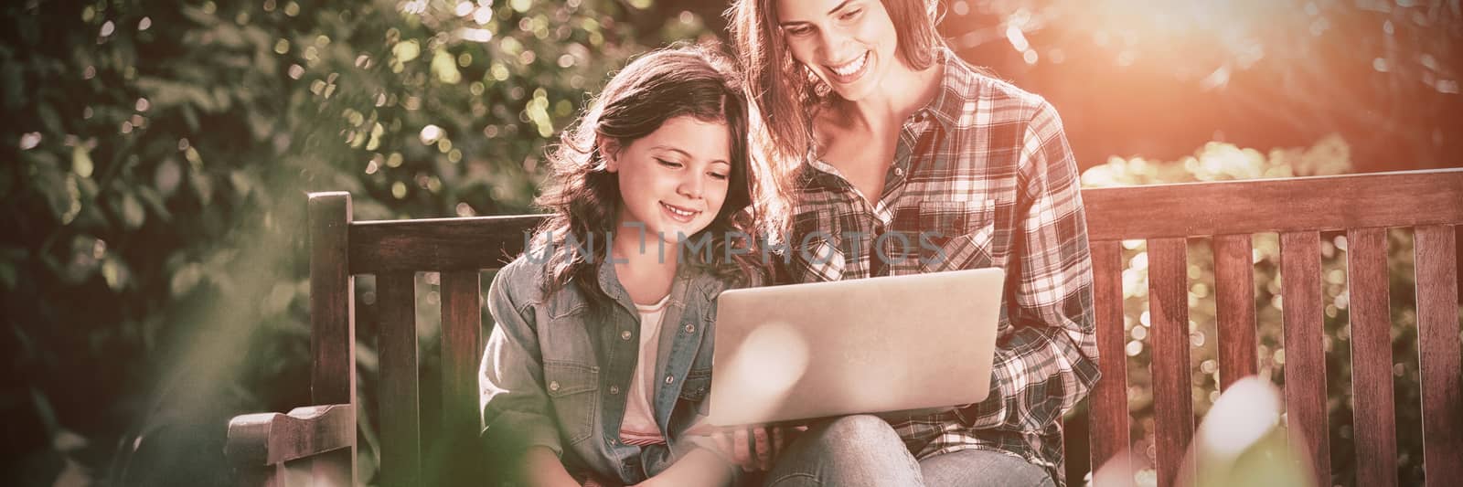 Smiling mother and daughter using laptop while sitting on wooden bench by Wavebreakmedia