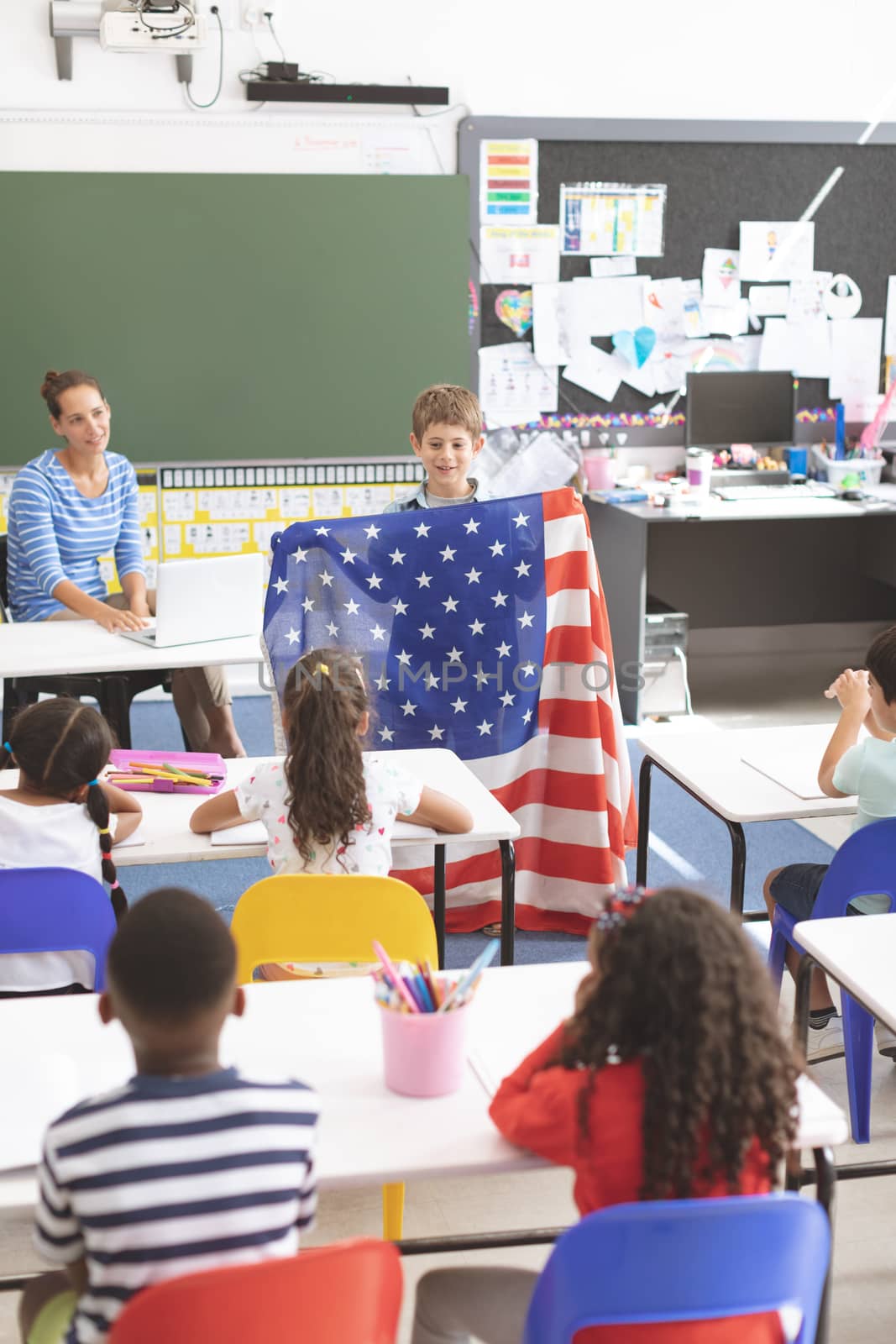 Front view of a schoolboy holding an american flag in classroom at school with his classmates in foreground