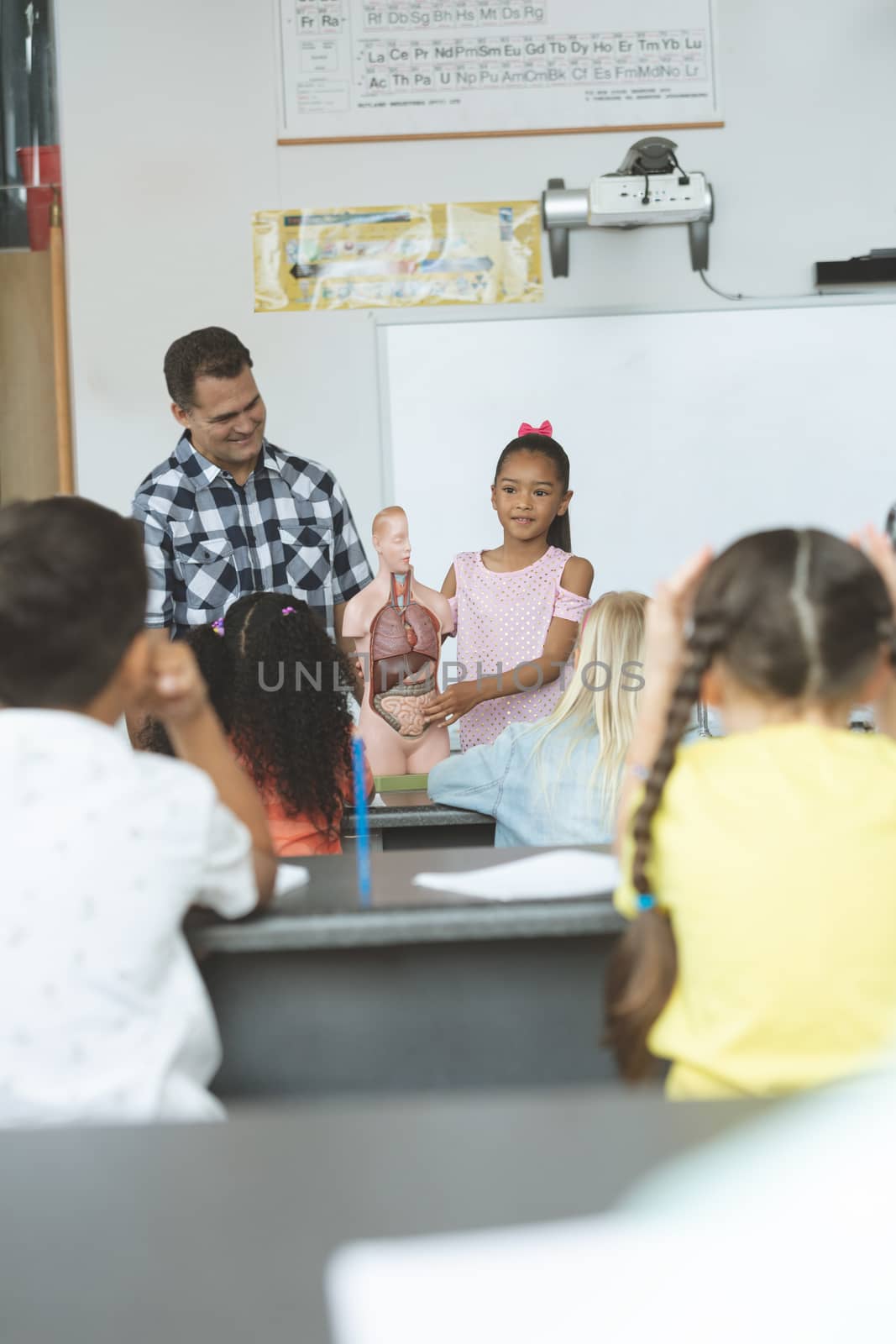 Front view of a schoolgirl explaining to his classmates the functioning of body human thanks to a dummy skeleton in classroom at school with his teacher next to her