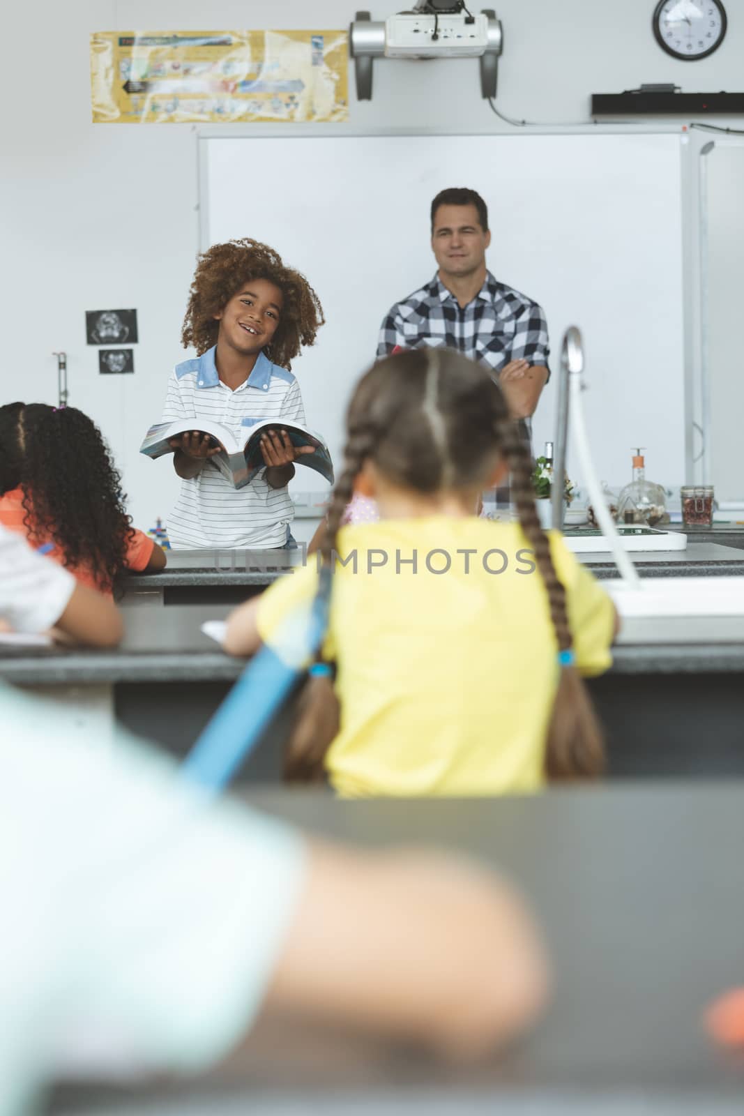 Front view of an African ethnicity schoolboy next to his teacher and holding a book  while looking at his classmates