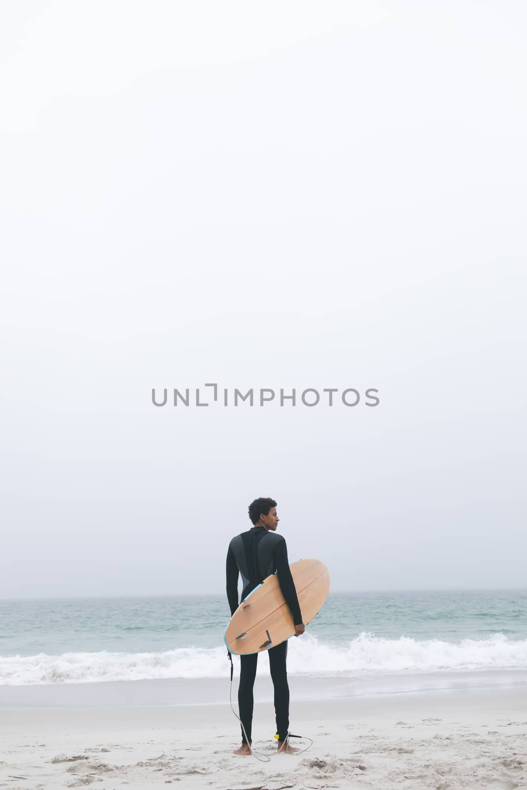 Rear view of thoughtful young mixed-race male surfer holding surfboard on the beach. He is looking away