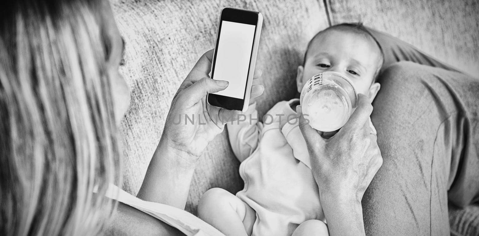 Mother using mobile phone while feeding her baby with milk bottle by Wavebreakmedia