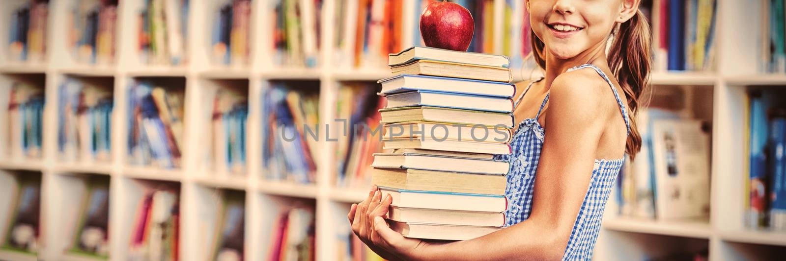 Portrait of school girl holding stack of books with apple in library at school