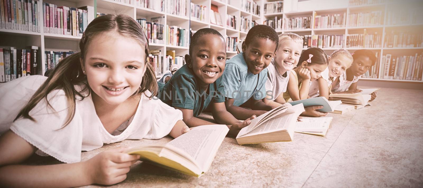 Smiling school kids lying on floor reading book in library  by Wavebreakmedia