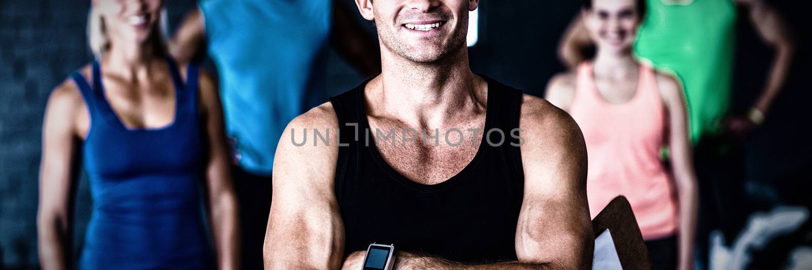 Portrait of smiling gym trainer holding clipboard while people standing in background