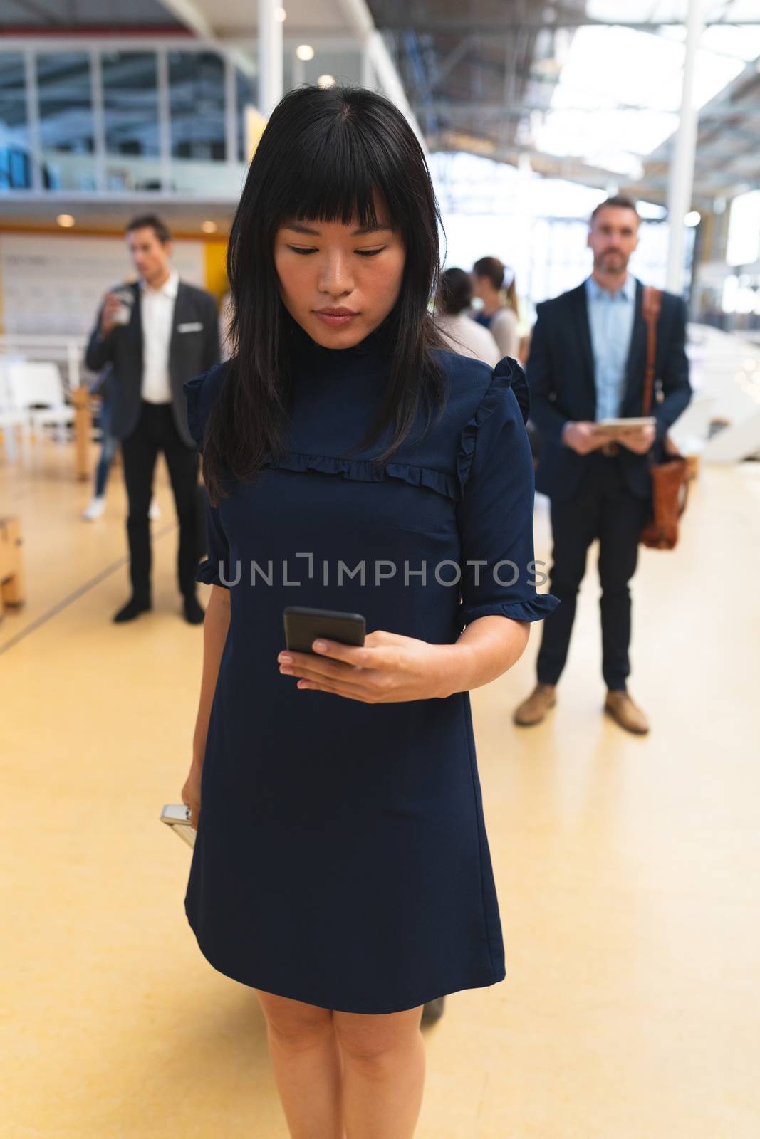 Close up of Asian businesswoman using mobile phone while standing in corridor at office with business people. International diverse corporate business partnership concept