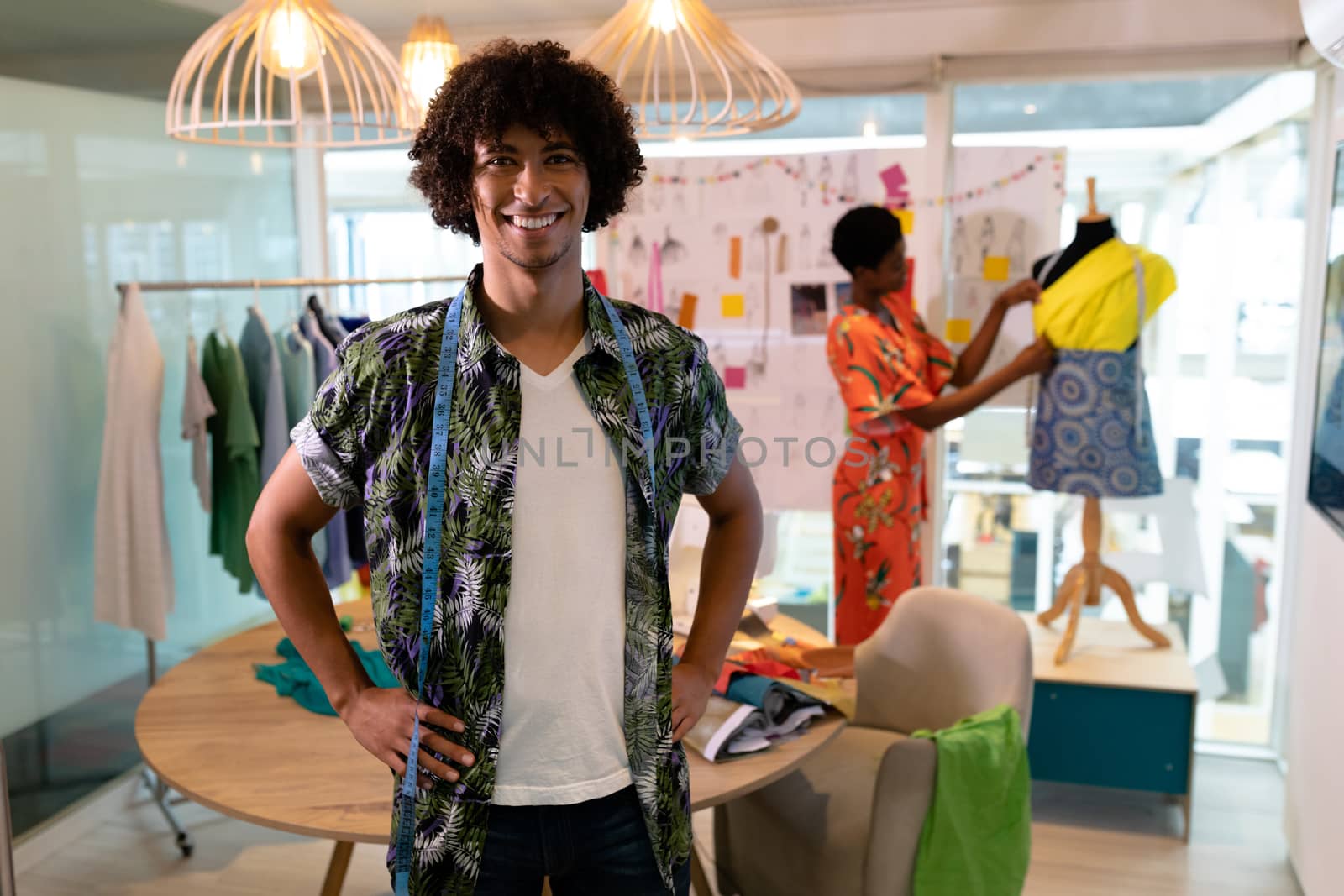 Portrait of young mixed race male fashion designer standing with hands on hip in design studio. African american woman working in the background. This is a casual creative start-up business office for a diverse team