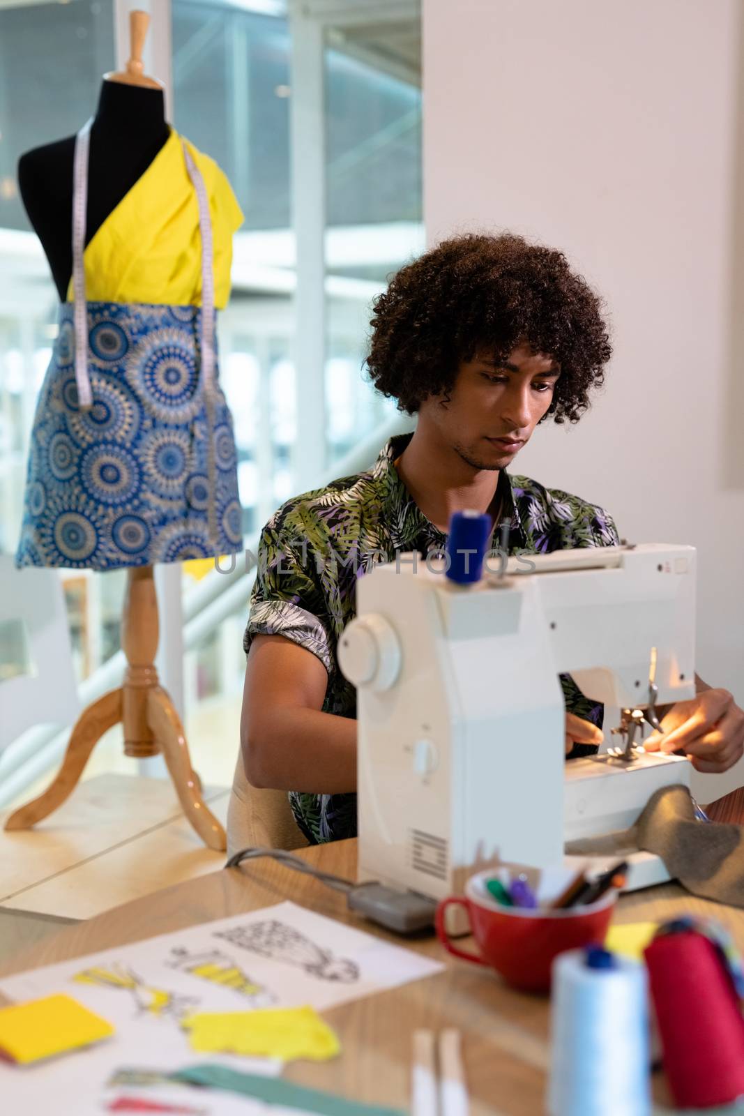 Front view of young mixed race male fashion designer using sewing machine on a table in design studio. This is a casual creative start-up business office for a diverse team