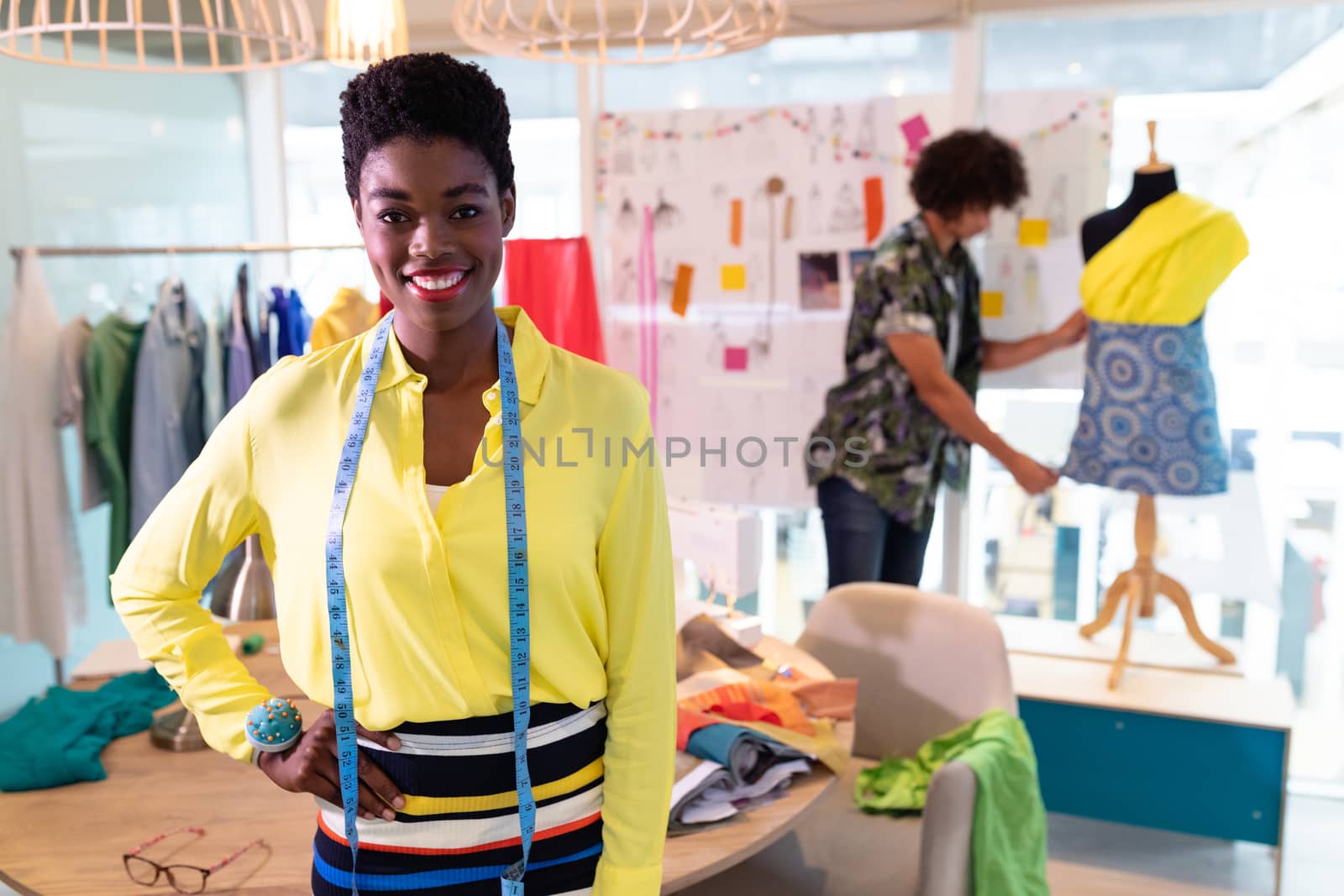 Front view of pretty young African american female fashion designer standing with hand on hip in design studio. Mixed race man working in the background. This is a casual creative start-up business office for a diverse team