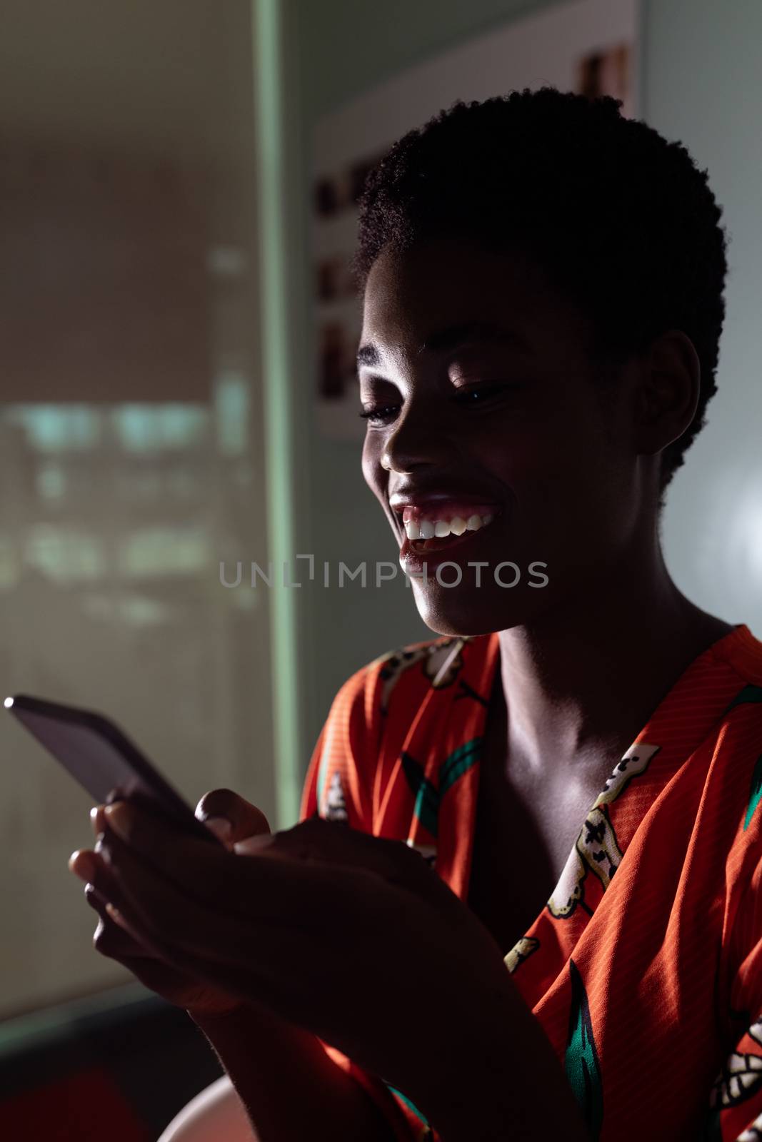 Side view of pretty young African american female graphic designer using mobile phone at desk in office. This is a casual creative start-up business office for a diverse team