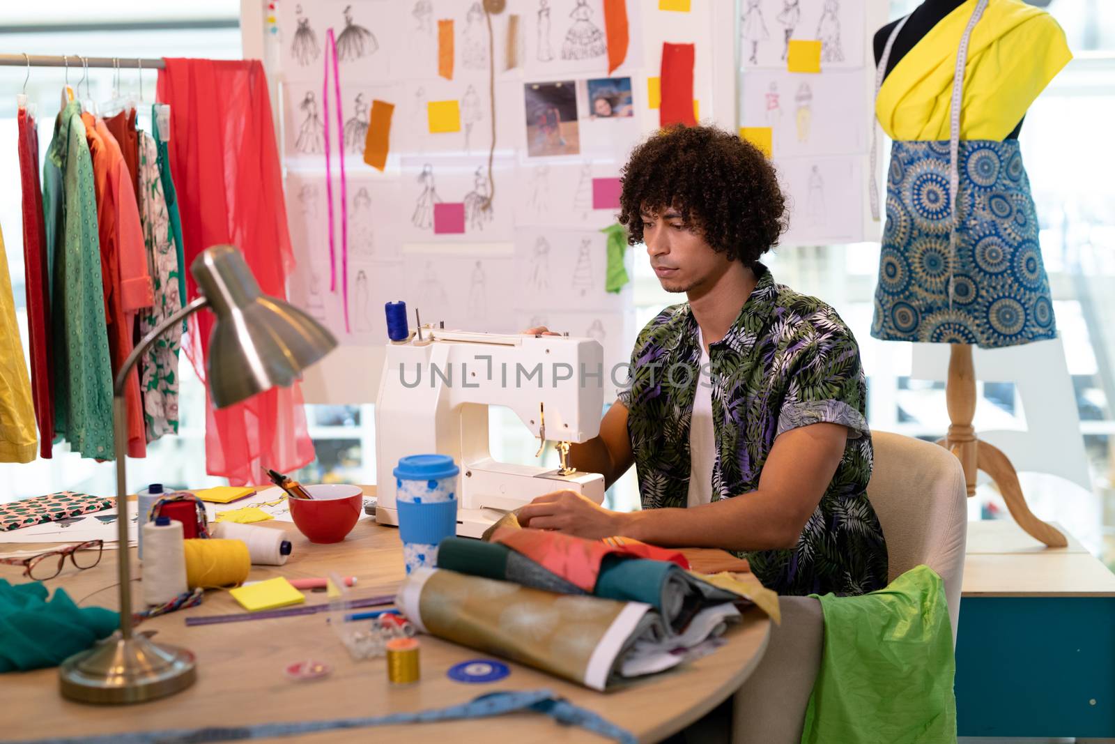 Side view of young mixed race male fashion designer using sewing machine on a table in design studio. This is a casual creative start-up business office for a diverse team