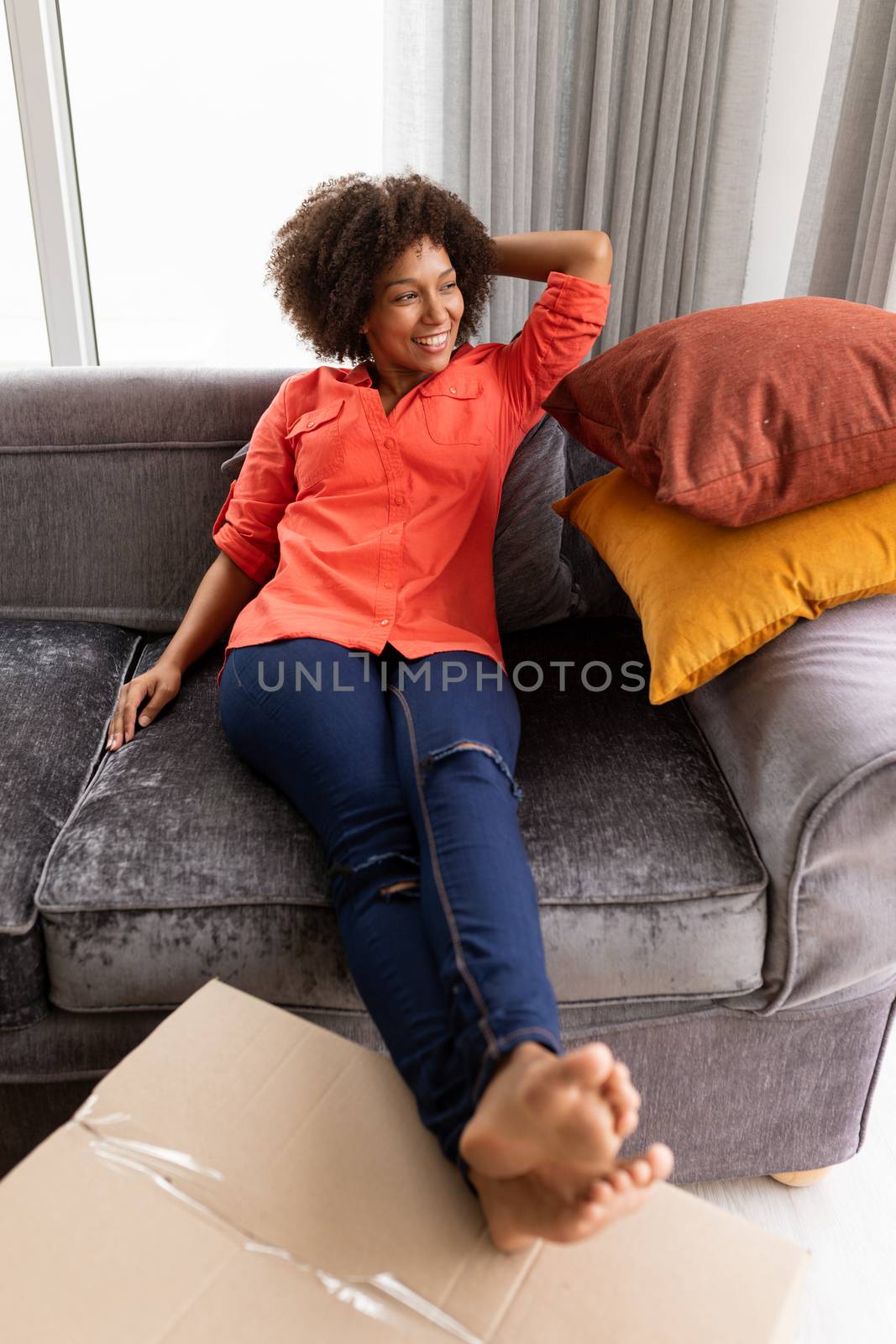 Front view of happy mixed-race woman relaxing on a sofa with her bare feet on the cardboard boxes in living room at home