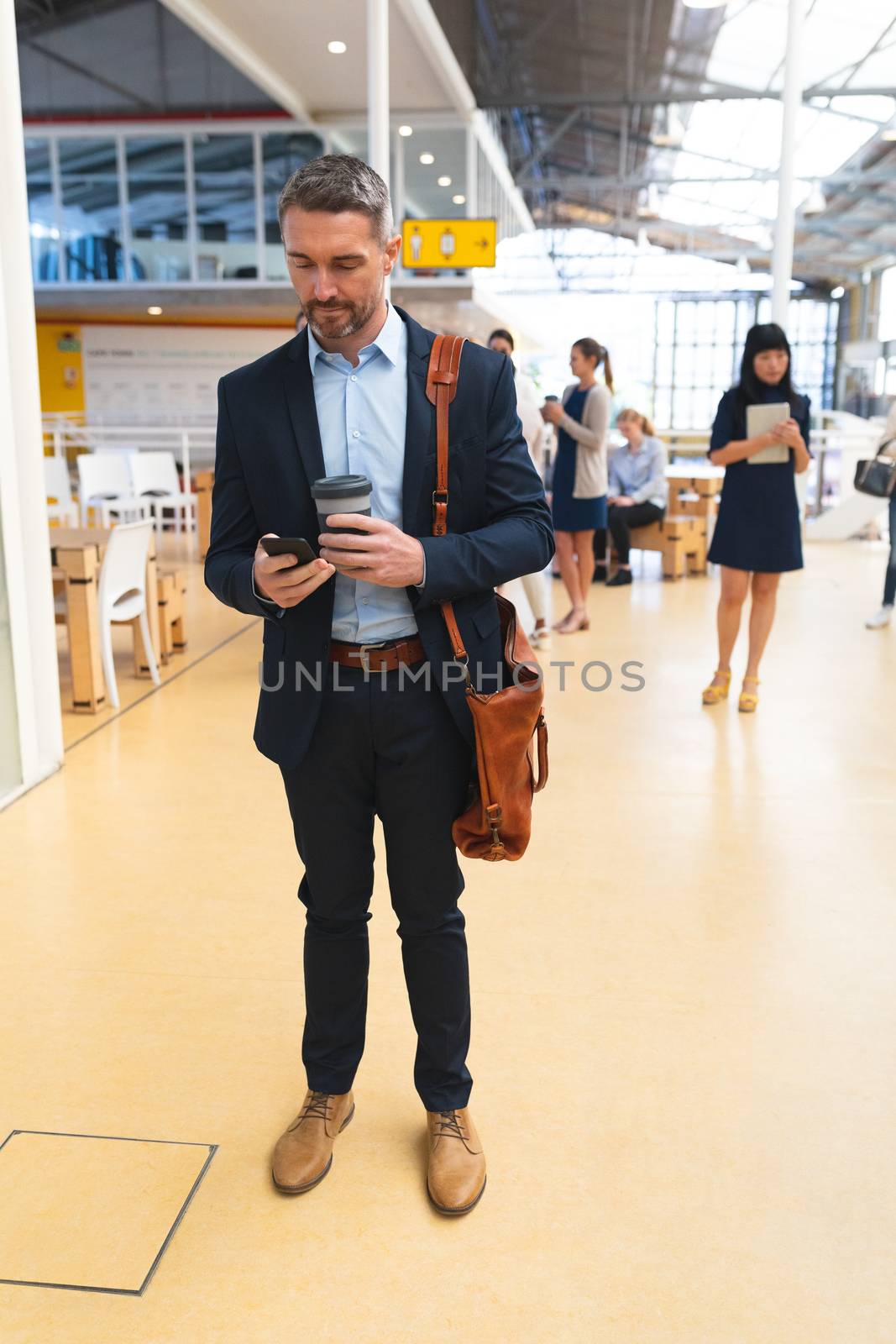 Front view of Caucasian businessman using mobile phone while standing in the corridor at office  with business people. International diverse corporate business partnership concept
