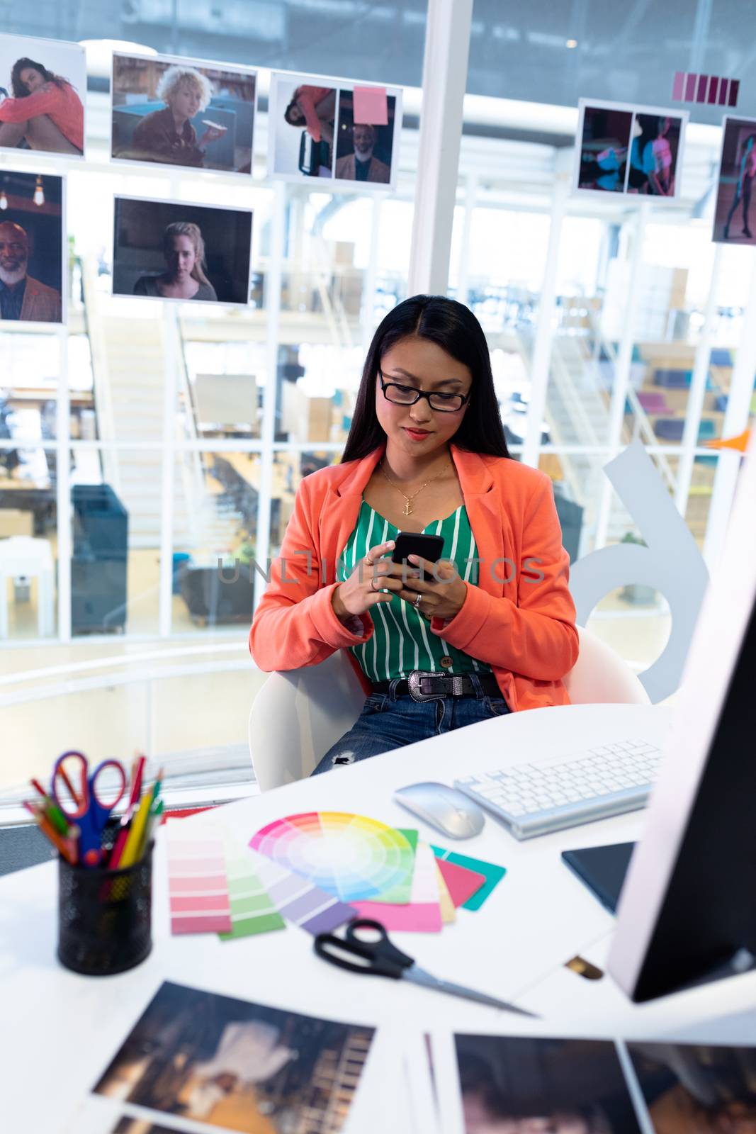 Front view of young pretty Asian female graphic designer using mobile phone at desk in office. This is a casual creative start-up business office for a diverse team