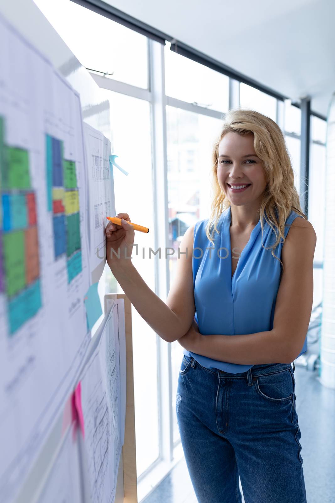 Front view of happy Caucasian female architect looking at camera while working over blueprint on whiteboard in a modern office. This is a casual creative start-up business office for a diverse team