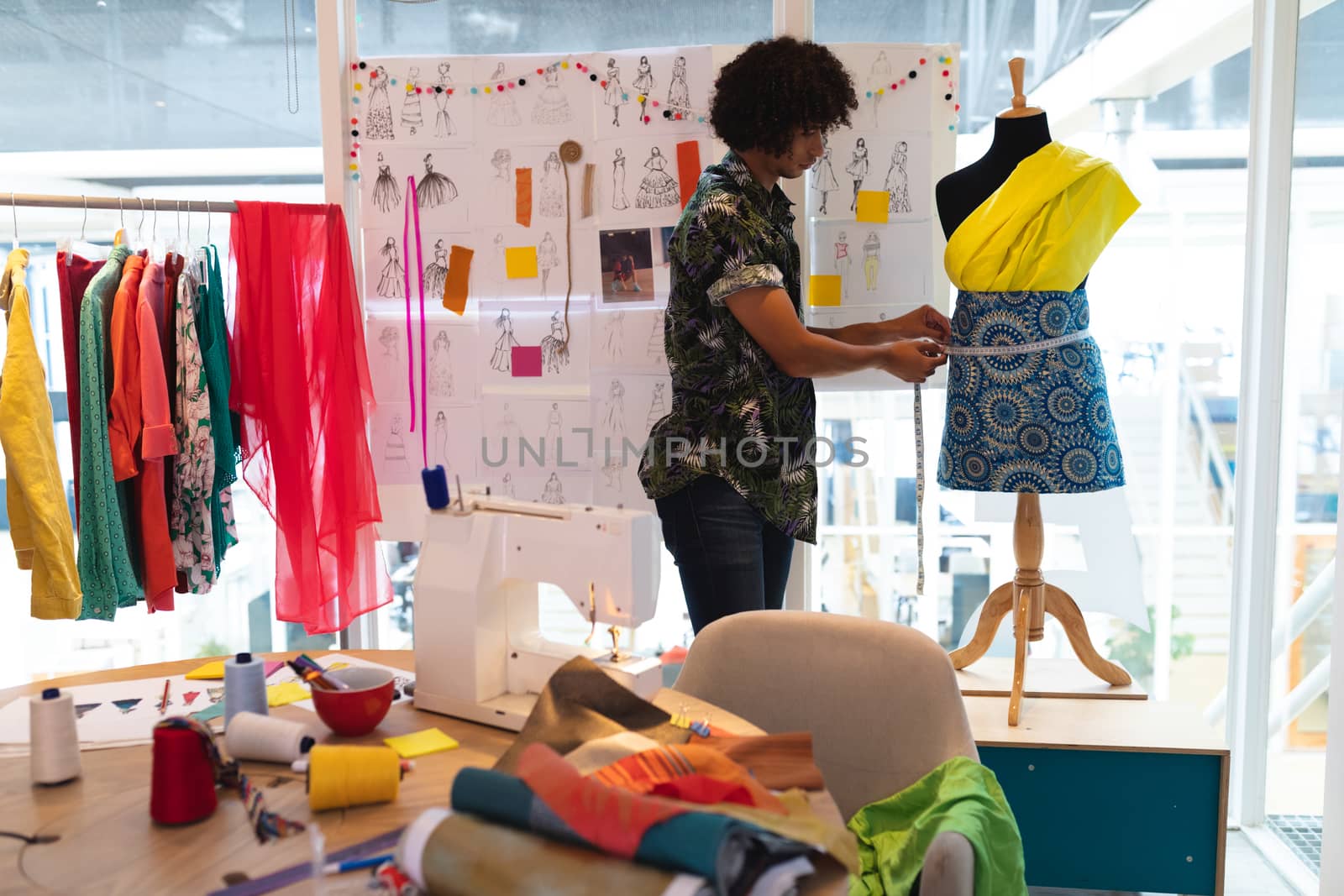 Side view of young mixed race male fashion designer using measuring tape on a mannequin in design studio. This is a casual creative start-up business office for a diverse team