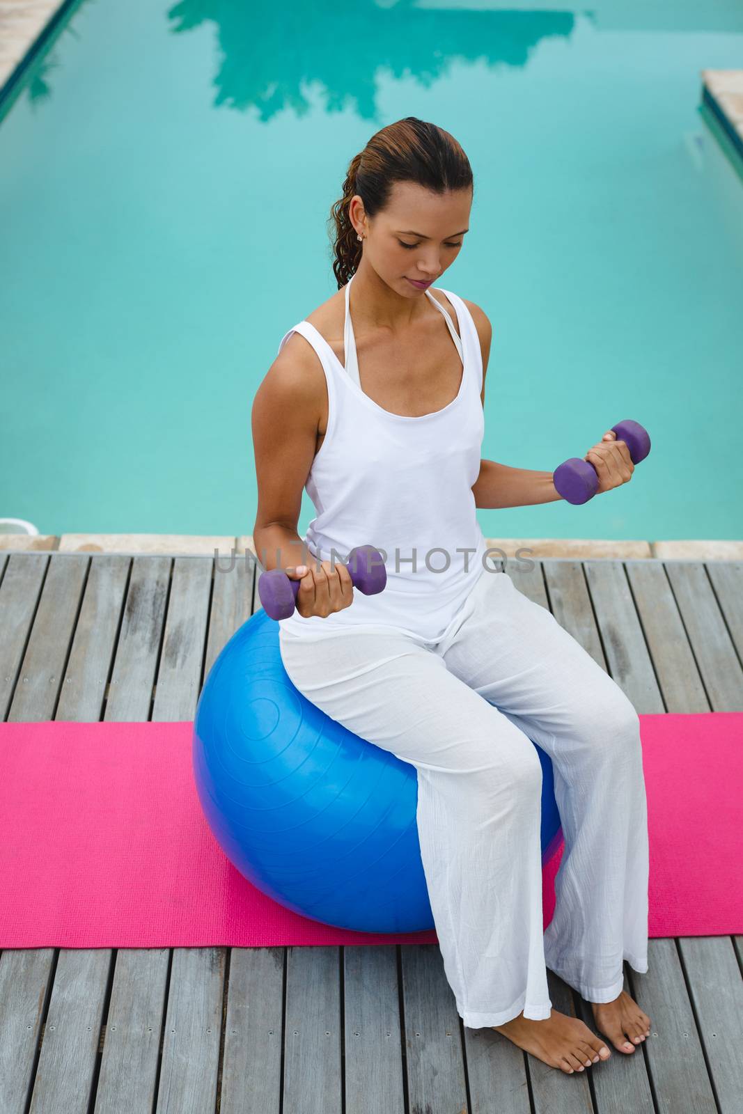 High angle view of fit mixed-race woman exercising with dumbbells while sitting on a exercise ball near swimming pool in the backyard. Summer fun at home by the swimming pool