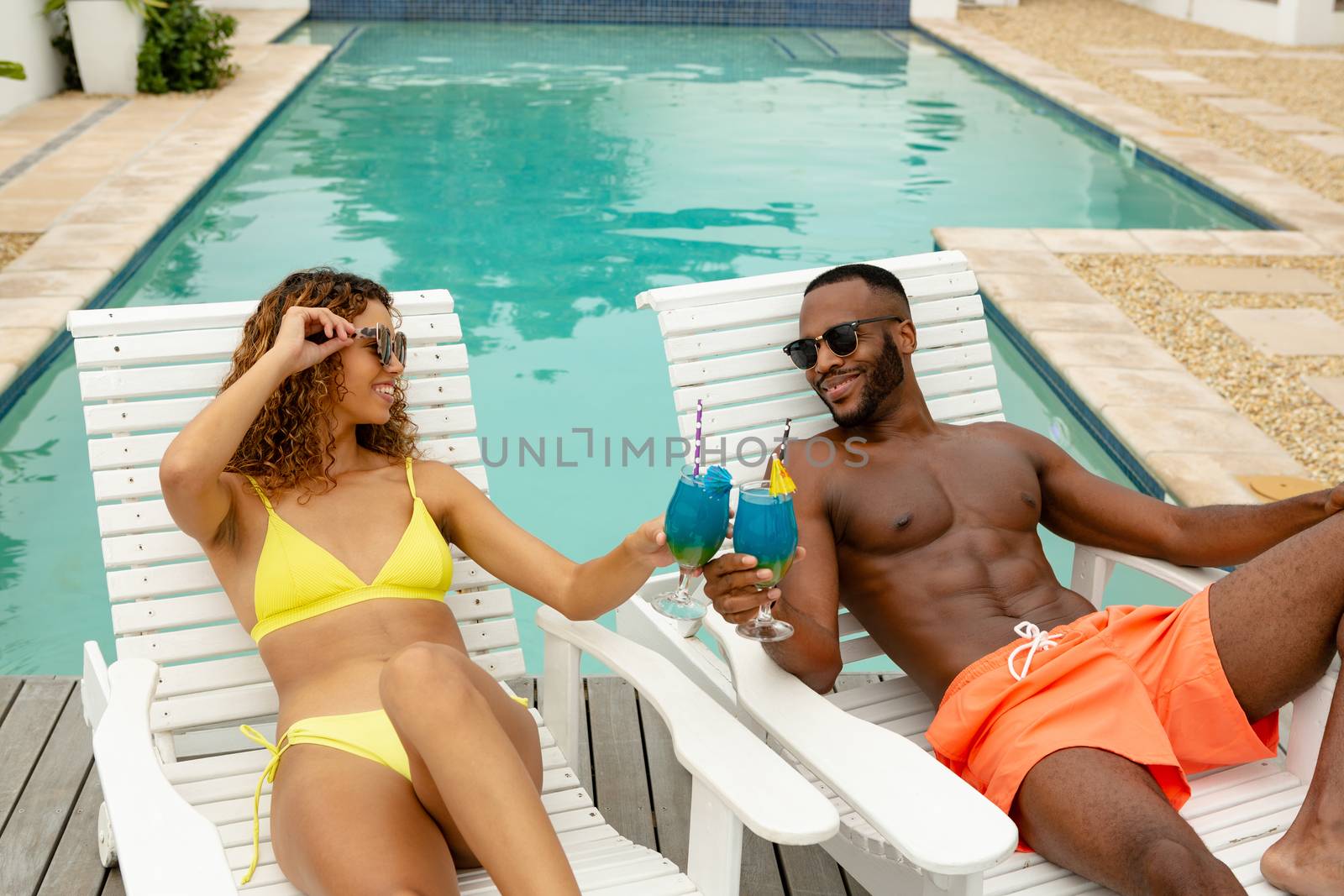 Front view of happy diverse couple toasting glasses of cocktail while relaxing on a sun lounger near swimming pool. Summer fun at home by the swimming pool