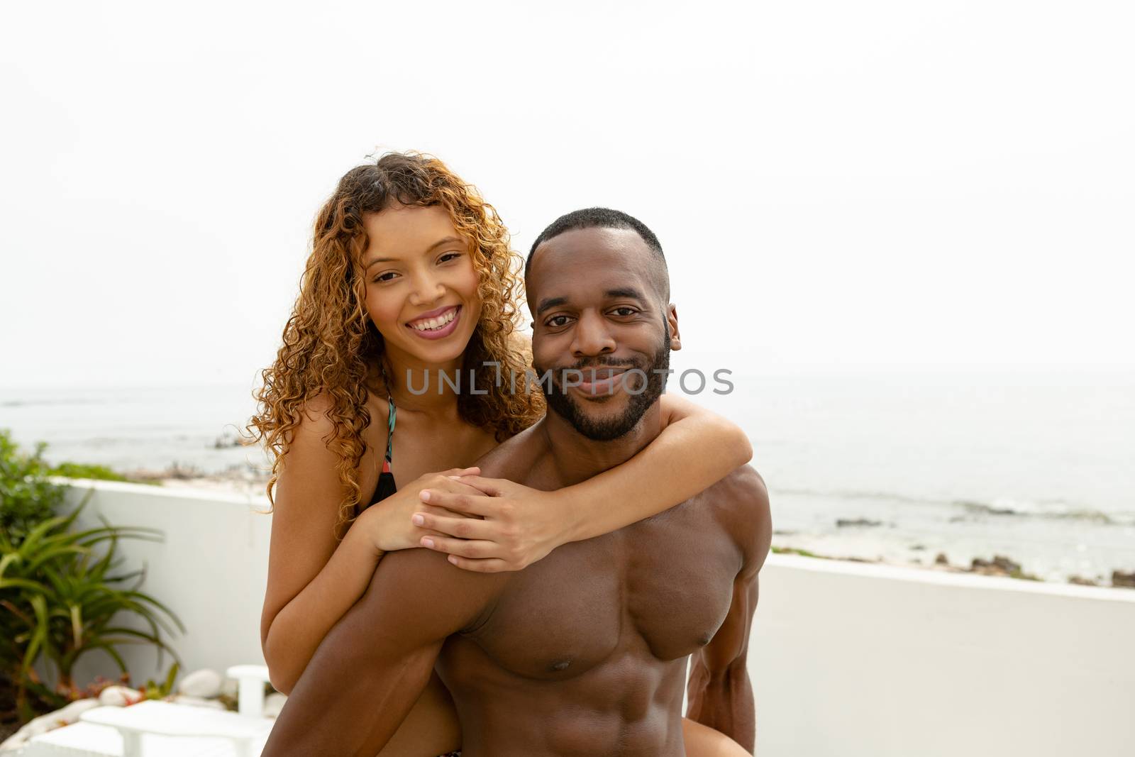 Portrait of African-american man giving piggyback ride to mixed-race woman near swimming pool at the backyard of home. Summer fun at home by the swimming pool
