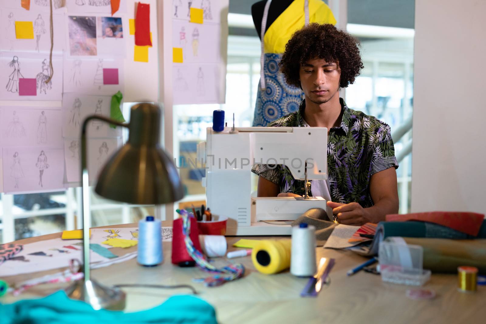 Front view of young mixed race male fashion designer using sewing machine on a table in design studio. This is a casual creative start-up business office for a diverse team