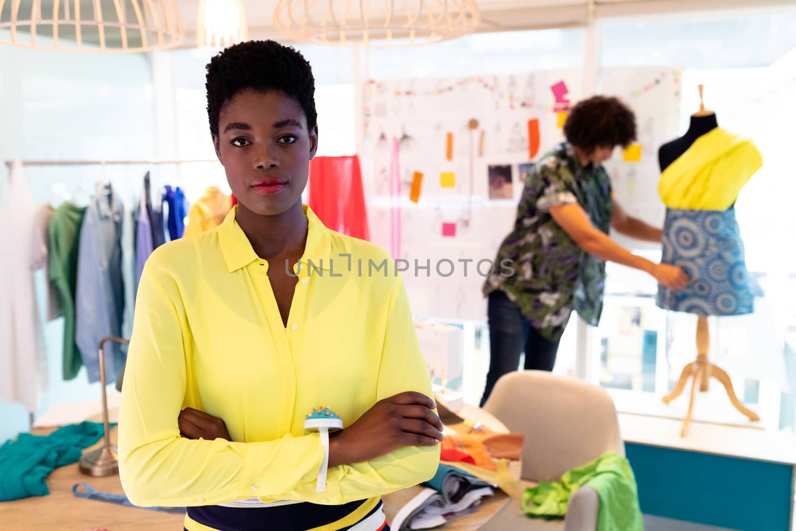 Front view of pretty young African american female fashion designer standing with arms crossed in design studio. Mixed race man working in the background. This is a casual creative start-up business office for a diverse team