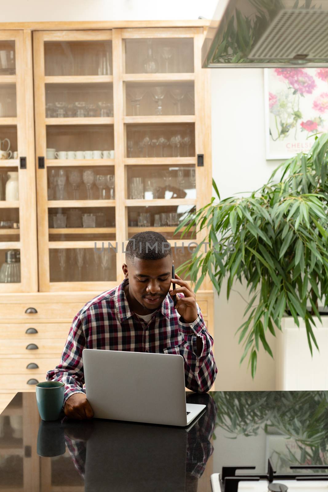 Front view of African-american man talking on mobile phone while using laptop on worktop in kitchen at comfortable home. Authentic home lifestyle setting with young African American male