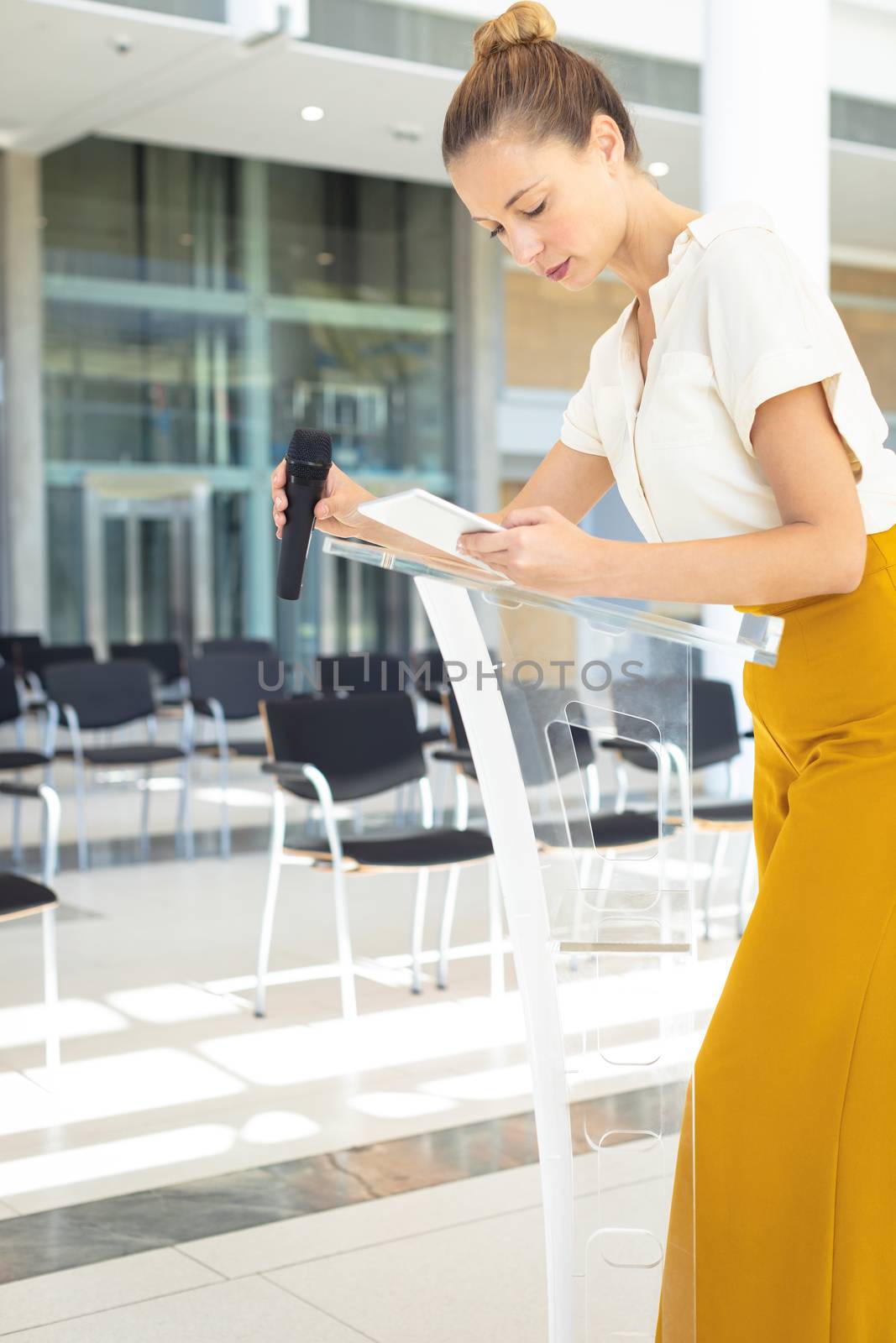 Side view of young Caucasian female executive standing with microphone while looking at digital tablet