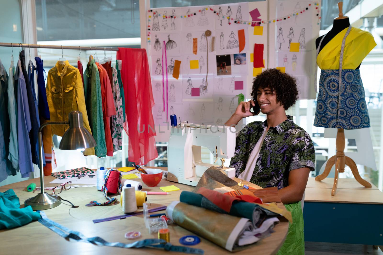 Side view of young mixed race male fashion designer talking on mobile phone while using sewing machine in design studio. This is a casual creative start-up business office for a diverse team