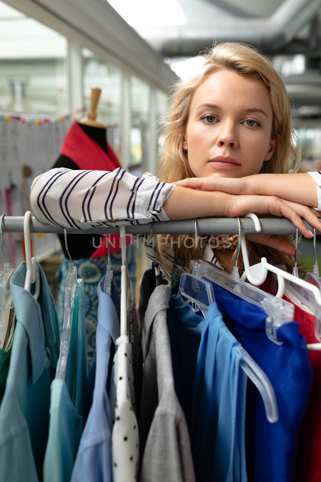 Portrait of young Caucasian female fashion designer leaning on clothes rack in design studio. This is a casual creative start-up business office for a diverse team